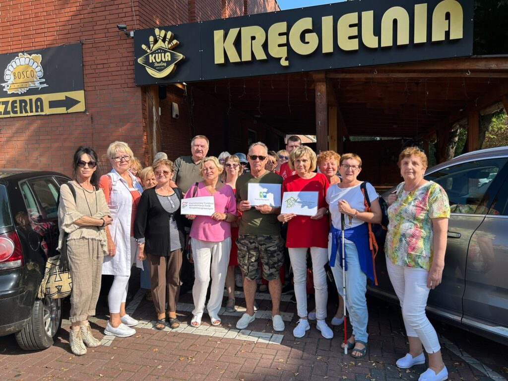 Grupa osób stoi przed ceglaną kręgielnią z drewnianym daszkiem; część trzyma kartki; obok widać szyld „PIZZERIA” ze strzałką w lewo i napis „KUŹNIA” nad wejściem; pod nogami brukowany chodnik i zaparkowane samochody.