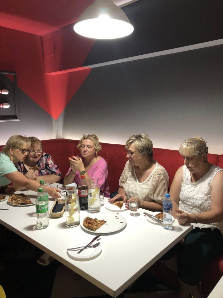 Sześć osób siedzi przy białym prostokątnym stole w restauracji z czerwonymi i szarymi ścianami; nad stołem wisi duża biała lampa; na stole leżą talerze z pizzą, butelki wody, butelka Sprite, szklanki z napojami i sztućce; uczestnicy rozmawiają i jedzą, tworząc swobodną, towarzyską atmosferę.