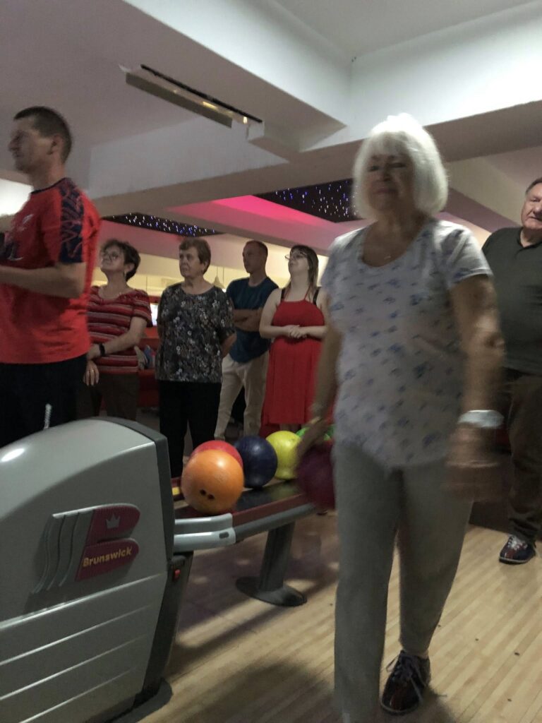 Kobieta w białej koszuli z niebieskim wzorem i szarych spodniach idzie w stronę kamery z różową kulą do kręgli; za nią stoją i rozmawiają uczestnicy przy torach, a na pierwszym planie widoczny jest zwrotnik Brunswick z kulami w kolorach pomarańczowym, fioletowym, zielonym i żółtym; sufit ma wbudowane oświetlenie i różowe akcenty.