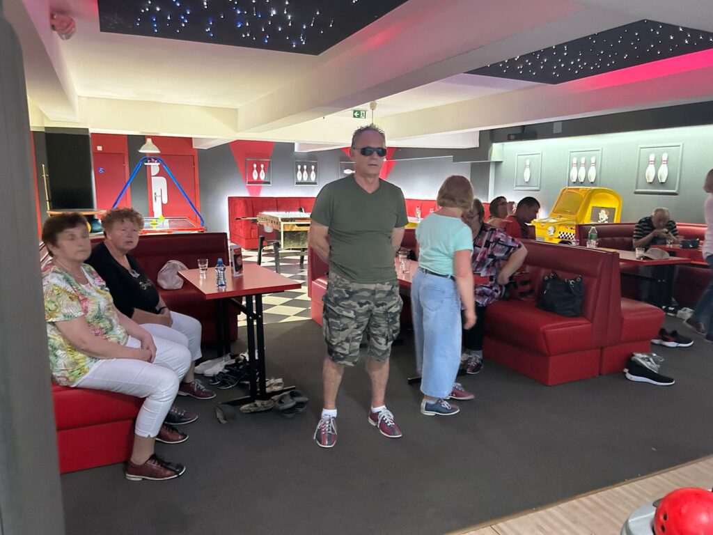 Grupa osób w kręgielni z czerwonymi siedzeniami i gwiaździstym sufitem; mężczyzna w zielonej koszulce, okularach przeciwsłonecznych i bojówkach stoi w centrum; obok widać kule i buty do kręgli.