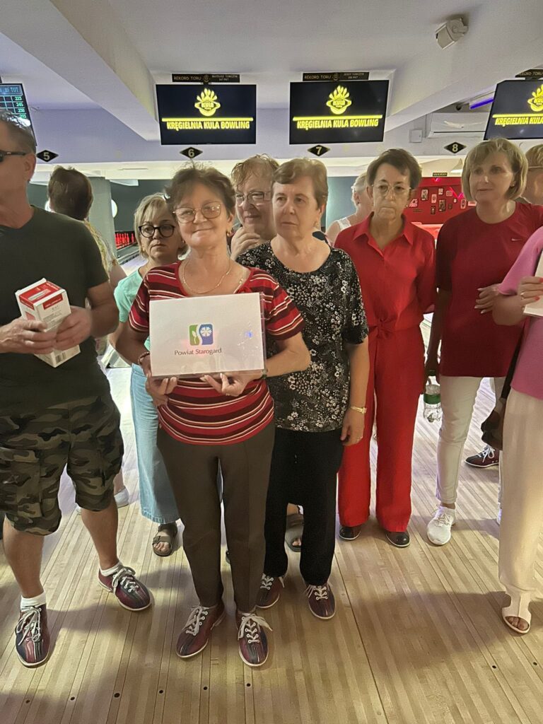 Grupa starszych osób stoi w kręgielni KULA BOWLING; niektórzy mają buty do kręgli, a kobieta z przodu trzyma biały karton z napisem Fundacja Śląskie Hospicjum dla Dzieci; w tle widać tory i ekrany z nazwą obiektu.