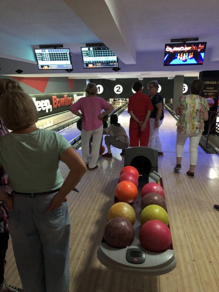 Grupa osób w czterotorowej kręgielni; na pierwszym planie wrzutnik kul z różowymi, pomarańczowymi, żółtymi i czerwonymi kulami; w tle ludzie stoją i siedzą przy torach oznaczonych numerami 1–4, nad nimi wiszą elektroniczne tablice wyników i ekrany; wnętrze jest przyciemnione, typowe dla kręgielni.