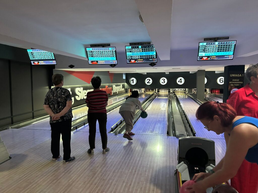 Grupa osób grająca w kręgle w sześciotorowej kręgielni; jedna osoba puszcza kulę na torze nr 3, a pozostali obserwują lub przygotowują się do rzutu; nad torami wiszą cyfrowe tablice wyników, wnętrze jest jasne i dobrze oświetlone.