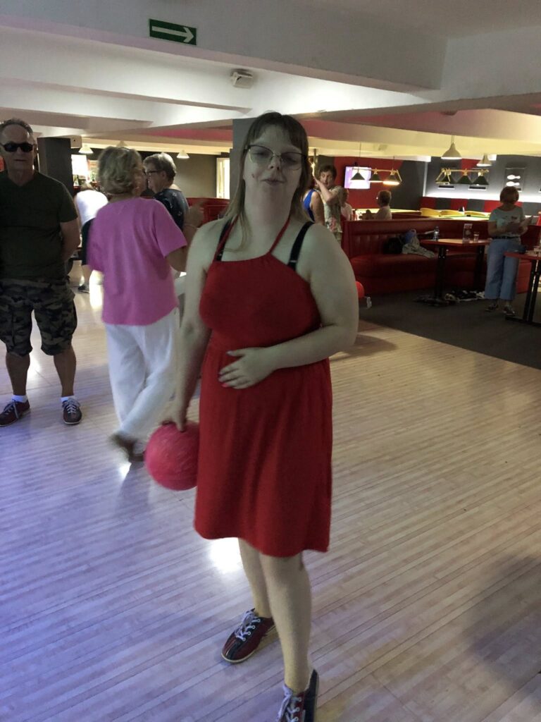 Osoba w czerwonej sukience i butach do kręgli stoi na drewnianym torze, trzymając różową kulę; w tle widać czerwone sofy, telewizory na ścianach oraz inne osoby siedzące lub stojące.