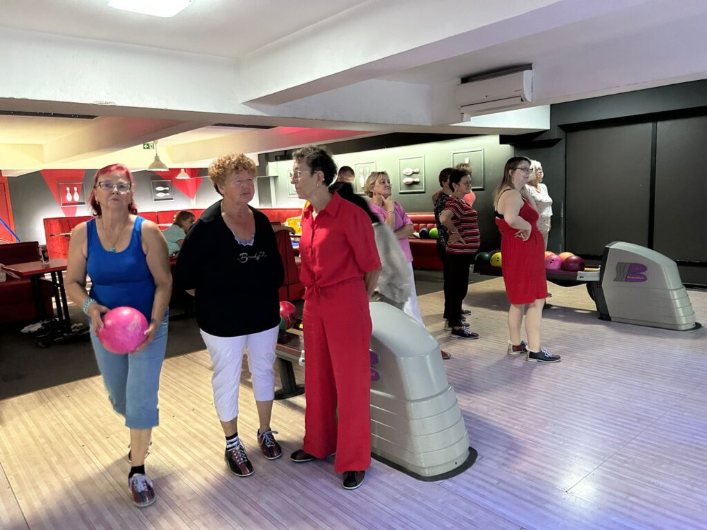 Grupa osób w nieformalnych ubraniach stoi przy torach do kręgli – niektórzy trzymają kule, inni rozmawiają; w tle widać czerwone sofy, różnokolorowe kule na powrocie, jasne drewniane tory i oświetlony biały sufit.