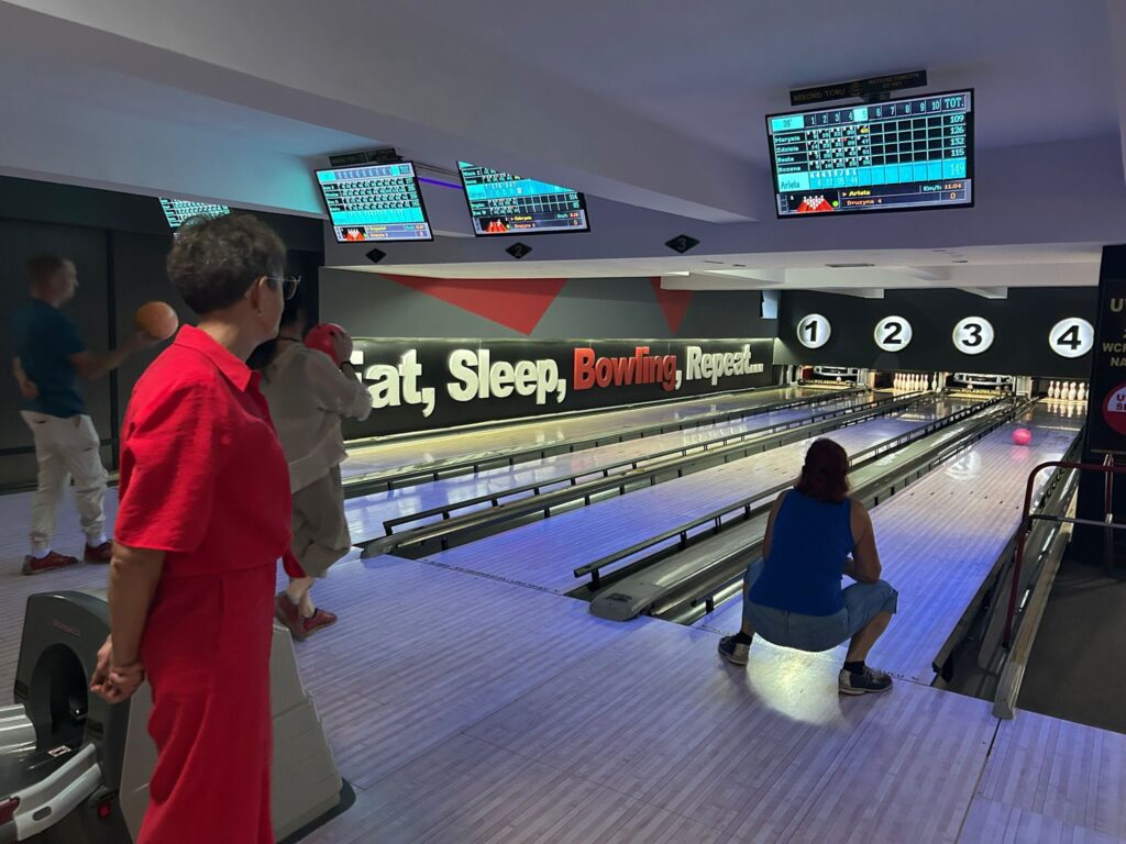 Trzecioplanowy widok czterotorowej kręgielni (numery torów 1–4) z kilkoma graczami: jedna osoba w czerwonym ubraniu stoi przy zwrocie kul, inna szykuje się do rzutu, a trzecia kuca przy torze; nad nimi elektroniczne tablice wyników, a na ścianie mural z napisem Eat, Sleep, Bowling, Repeat… ze słowem Bowling wyróżnionym na czerwono.