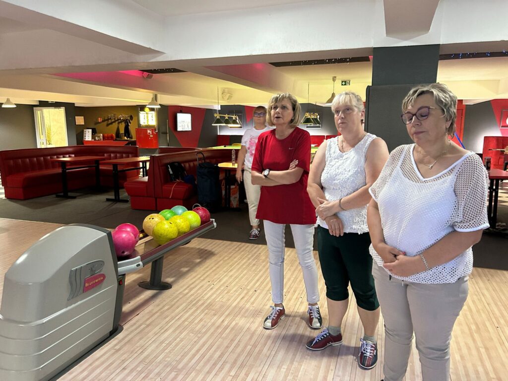 Trzy osoby w nieformalnych ubraniach i butach do kręgli stoją przy urządzeniu do zwrotu kul w kręgielni, obok różowych, żółtych i zielonych kul; w tle widać czerwone siedzenia, ladę i drewniane tory.