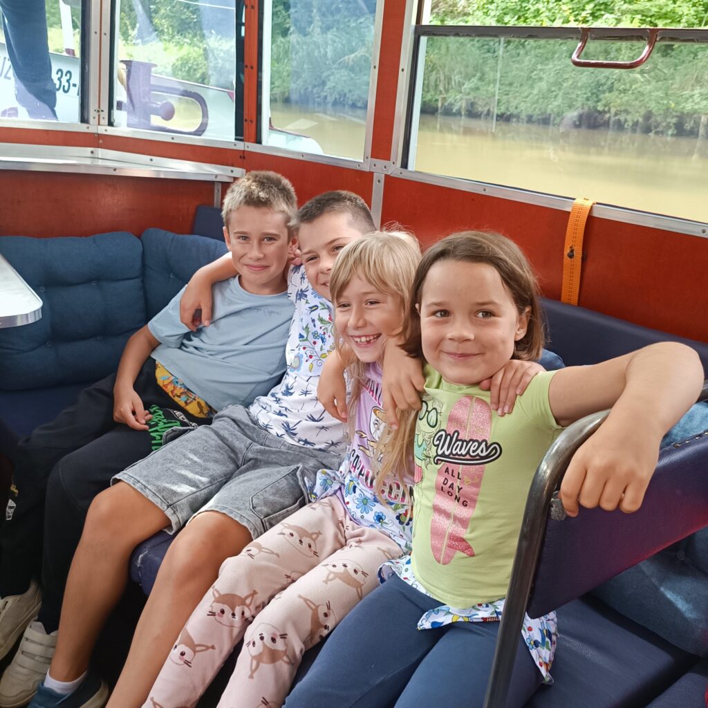 Czwórka uśmiechniętych dzieci siedzi razem na kanapie wewnątrz łodzi płynącej po Kanale Elbląskim, obejmując się ramionami. Za nimi duże okna z widokiem na brązową wodę kanału i zielony brzeg. Wnętrze wykończone drewnem, niebieskimi poduszkami i metalowymi detalami. Panuje radosna, przyjacielska atmosfera wycieczki.