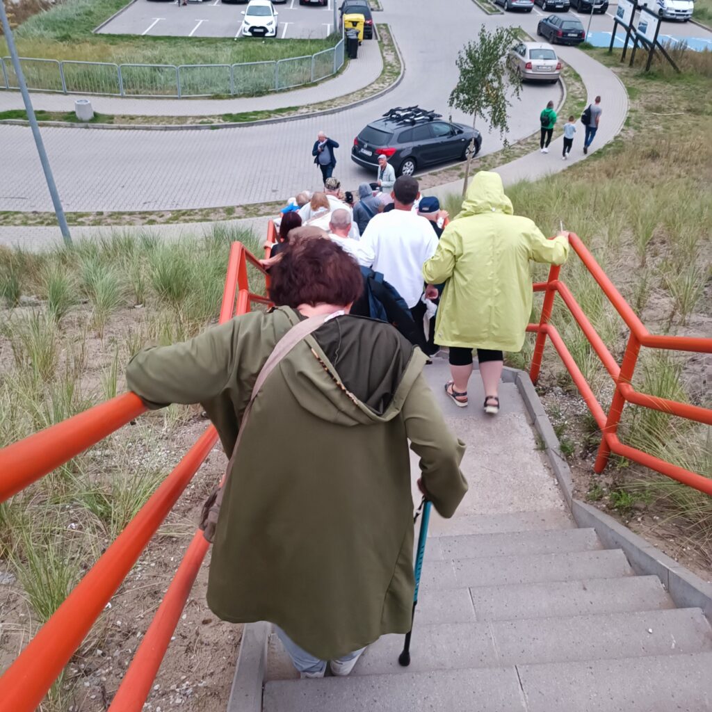 Grupa osób schodzi po betonowych schodach z pomarańczowymi poręczami. Jedna osoba korzysta z laski ortopedycznej. Uczestnicy ubrani w zielone i oliwkowe kurtki. Po bokach trawa, na dole chodnik i parking z samochodami. Pogoda chłodna lub deszczowa.