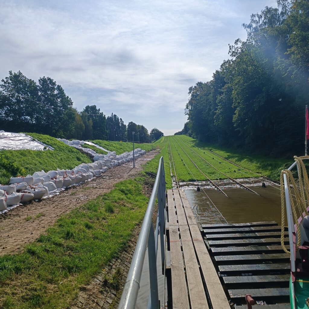 Drewniany pomost prowadzący do kanału wodnego oraz pochyłość z metalowymi szynami do transportu łodzi. Po prawej zalesiona skarpa, po lewej worki z piaskiem częściowo przykryte folią. Otoczenie zielone, w tle drzewa i niebieskie niebo z lekkim zachmurzeniem.