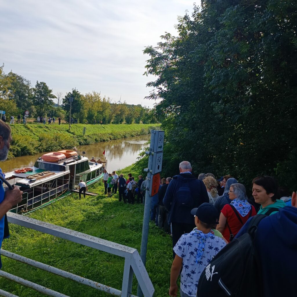 Uczestnicy wyjazdu rehabilitacyjnego ustawieni w kolejce na trawiastym brzegu kanału, czekają na wejście na zacumowany statek pasażerski. W tle spokojna woda, drzewa i metalowa barierka wyznaczająca ścieżkę dojścia. Zdjęcie oddaje atmosferę oczekiwania i wspólnego przeżywania wycieczki.