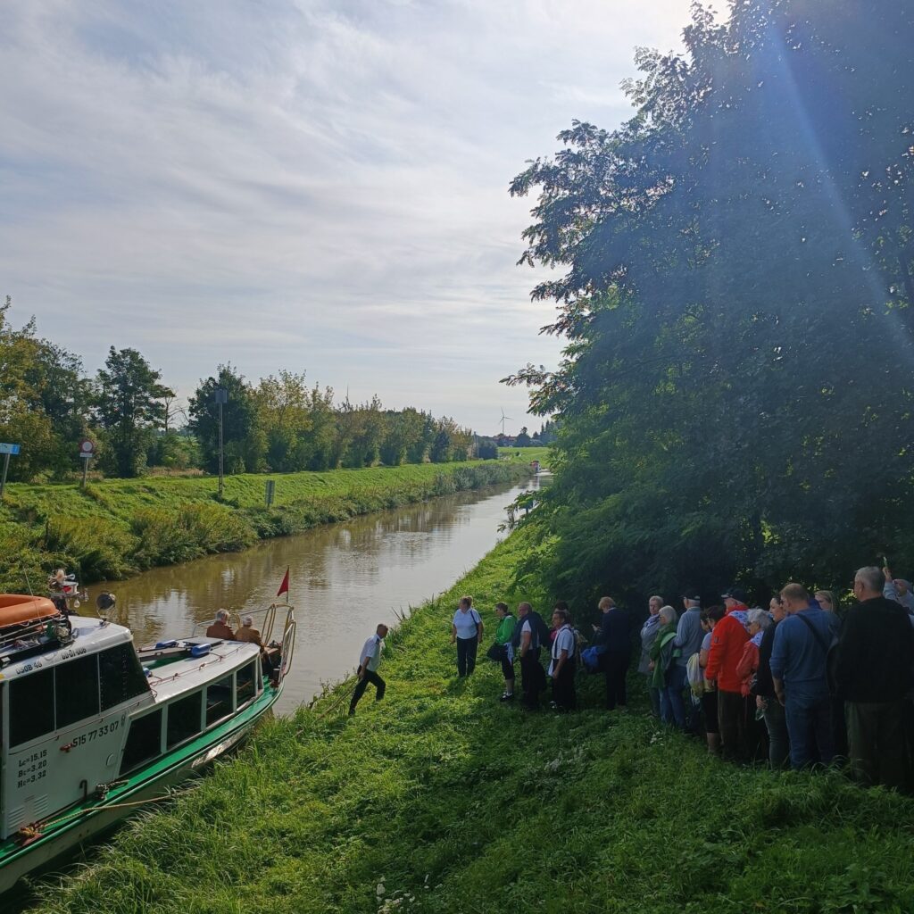 Grupa uczestników wyjazdu rehabilitacyjnego stoi na trawiastym brzegu kanału, obok zacumowanego zielono-białego statku. Jedna osoba schodzi z pokładu na brzeg. W tle spokojna woda, drzewa po obu stronach kanału i bezchmurne niebo. Zdjęcie ukazuje moment zakończenia rejsu i wspólnego przeżywania wycieczki w naturalnym otoczeniu.