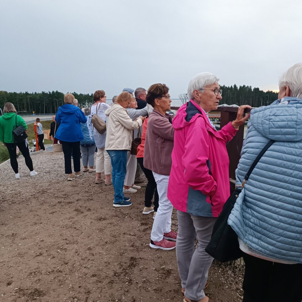 Grupa starszych uczestników wyjazdu rehabilitacyjnego stoi na żwirowej ścieżce w otoczeniu lasu i otwartego pola. Część osób opiera się o barierkę, patrząc w dal. Ubrani w kurtki, co sugeruje chłodniejszą pogodę. Niebo zachmurzone, atmosfera spokojna i kontemplacyjna.