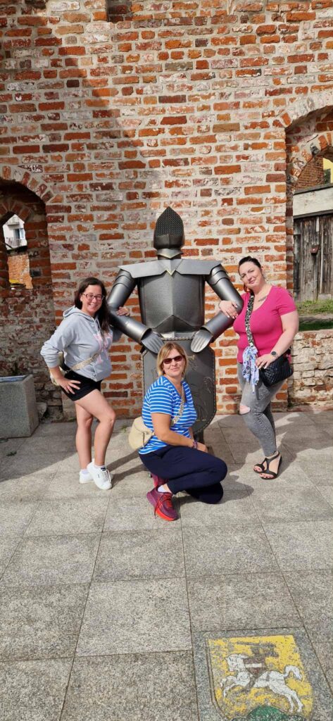 Trzy osoby pozują przy metalowej rzeźbie rycerza w zbroi, ustawionej na tle ceglanego muru z łukowymi wnękami. Jedna osoba klęczy z przodu, dwie stoją po bokach figury. W tle widoczna kolorowa mozaika lub herb na brukowanym placu. Atmosfera radosna, turystyczna, z nutą historyczną.