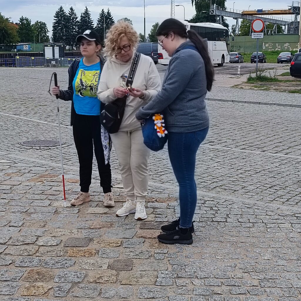Trzy osoby stoją na brukowanym placu w pobliżu autobusu i zielonego muralu. Jedna z nich, ubrana w niebieską koszulkę z żółtym nadrukiem, czapkę i czarne spodnie, trzyma białą laskę sygnalizującą niepełnosprawność wzroku. Druga osoba w jasnym ubraniu patrzy na telefon, trzecia ma szarą bluzę, dżinsy i niebieską torbę z brelokiem. W tle znaki drogowe i trawiasty teren. Atmosfera codzienna, komunikacyjna.