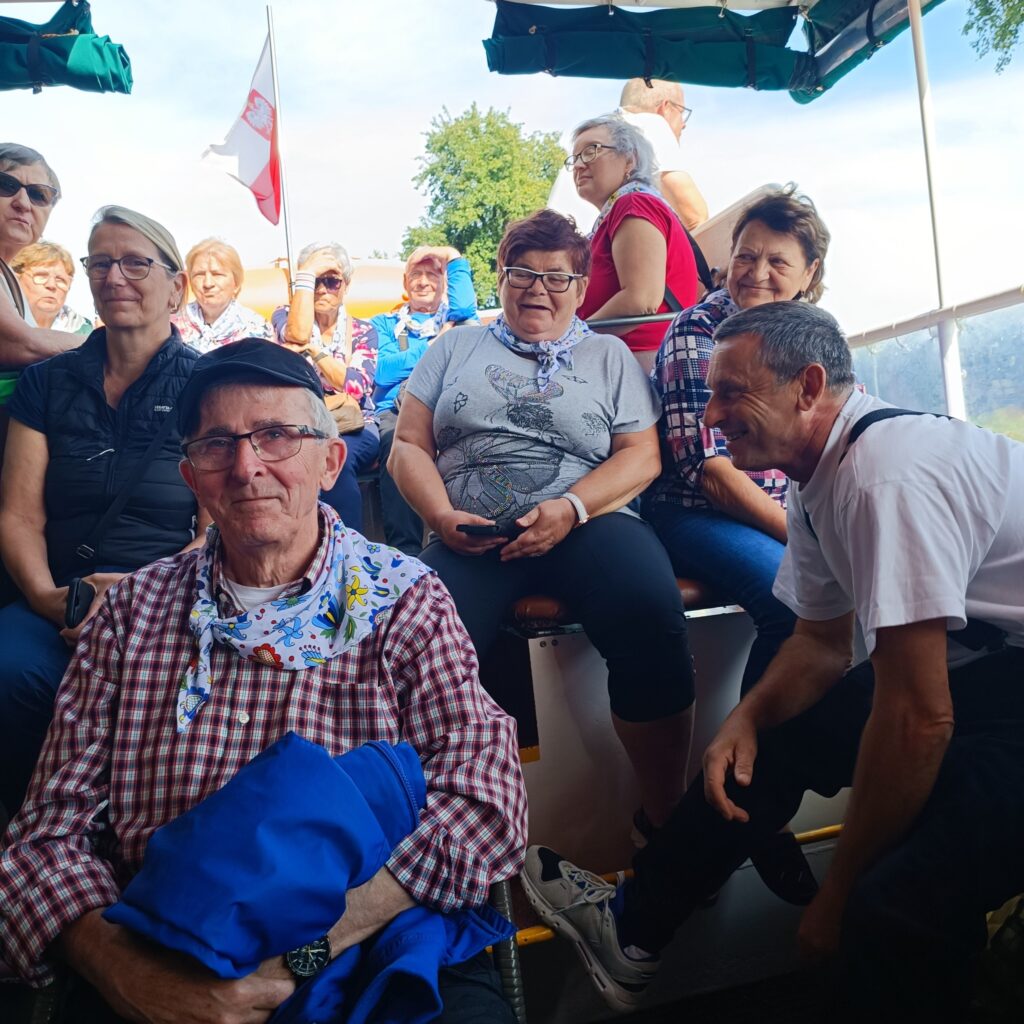 Grupa starszych uczestników wyjazdu rehabilitacyjnego siedzi blisko siebie na pokładzie statku. Część osób ma okulary przeciwsłoneczne, ubrania codzienne i pogodny wyraz twarzy. W tle widoczna flaga Polski, drzewa i parasole. Atmosfera towarzyska, słoneczna i wypoczynkowa.