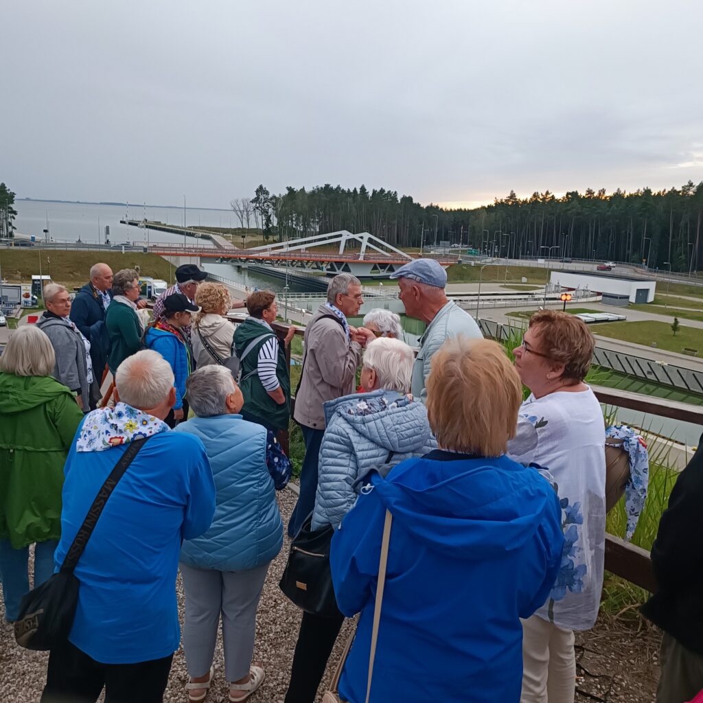 Grupa starszych uczestników wyjazdu rehabilitacyjnego stoi przy nowoczesnym białym moście łukowym nad kanałem lub rzeką. Osoby ubrane w kurtki, część słucha przewodnika. W tle drzewa, woda i uporządkowana przestrzeń rekreacyjna. Atmosfera edukacyjna i spokojna.