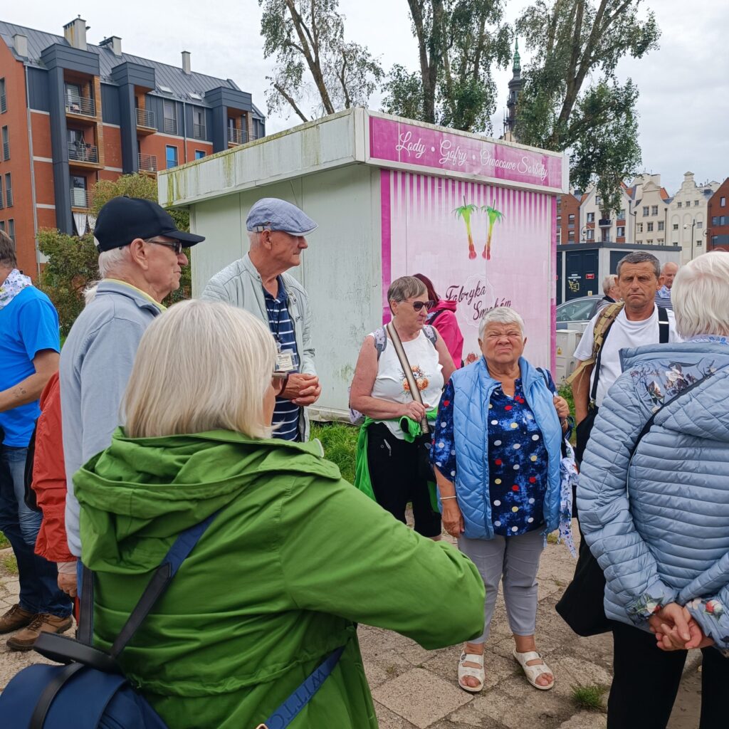 Grupa starszych uczestników wyjazdu rehabilitacyjnego stoi przy kolorowym kiosku z napisem „Lody • Gofry • Shake • Smoothie” oraz „Lody Trzy Świnki”. Osoby ubrane w kurtki i czapki, pogoda chłodna. W tle nowoczesne bloki mieszkalne i drzewa. Atmosfera towarzyska i pogodna.