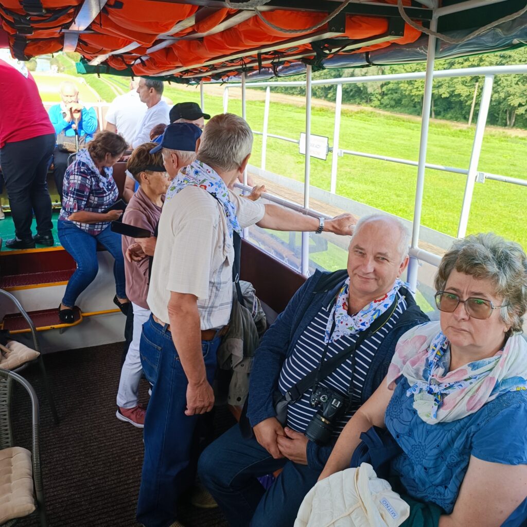 Grupa uczestników wyjazdu rehabilitacyjnego siedzi i stoi na pokładzie zadaszonego statku z przezroczystymi panelami bocznymi. Nad głowami widoczne pomarańczowe kamizelki ratunkowe. Osoby ubrane w codzienne stroje, część ma niebiesko-białe apaszki. W tle zielona trawa i drzewa. Atmosfera wycieczkowa, spokojna i towarzyska.