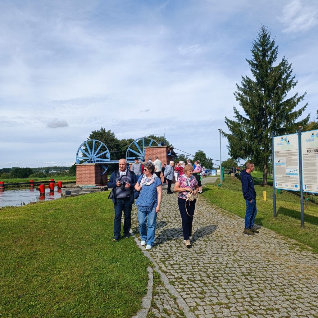 Grupa uczestników wyjazdu rehabilitacyjnego spaceruje brukowaną ścieżką obok dwóch dużych niebieskich kół wodnych zamontowanych na ceglanych konstrukcjach. W tle trawa, drzewa i tablice informacyjne z napisem „Kanał Elbląski”. Widoczna duża sosna po prawej stronie. Pogoda częściowo pochmurna, atmosfera edukacyjna i spokojna.