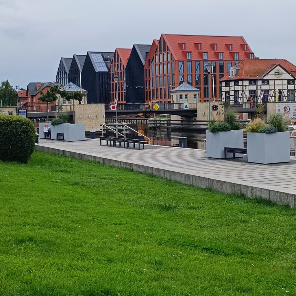 Widok na zadbane nadbrzeże z zielonym trawnikiem, drewnianym deptakiem z ławkami i donicami z ozdobnymi trawami. Po drugiej stronie kanału nowoczesne budynki z czerwonymi i czarnymi dachami, duże okna i styl łączący tradycję z nowoczesnością. Widoczny mały mostek nad wodą, spacerujący ludzie i spokojna atmosfera.