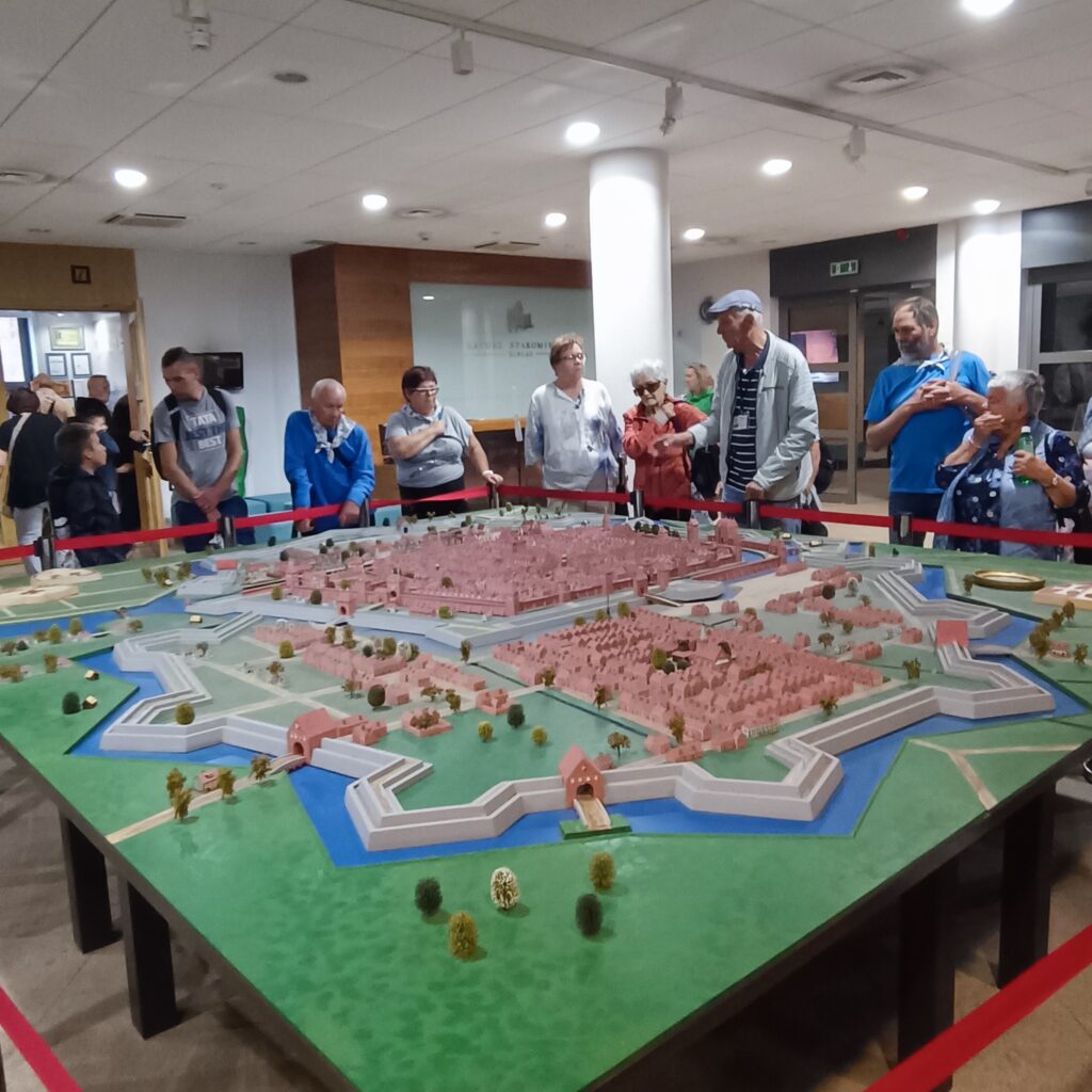 Grupa starszych uczestników wyjazdu rehabilitacyjnego stoi przy kolorowym kiosku z napisem „Lody • Gofry • Shake • Smoothie”. Osoby ubrane w kurtki i czapki, otoczenie miejskie z nowoczesnymi blokami i drzewami. Atmosfera towarzyska, pogodna i relaksująca.