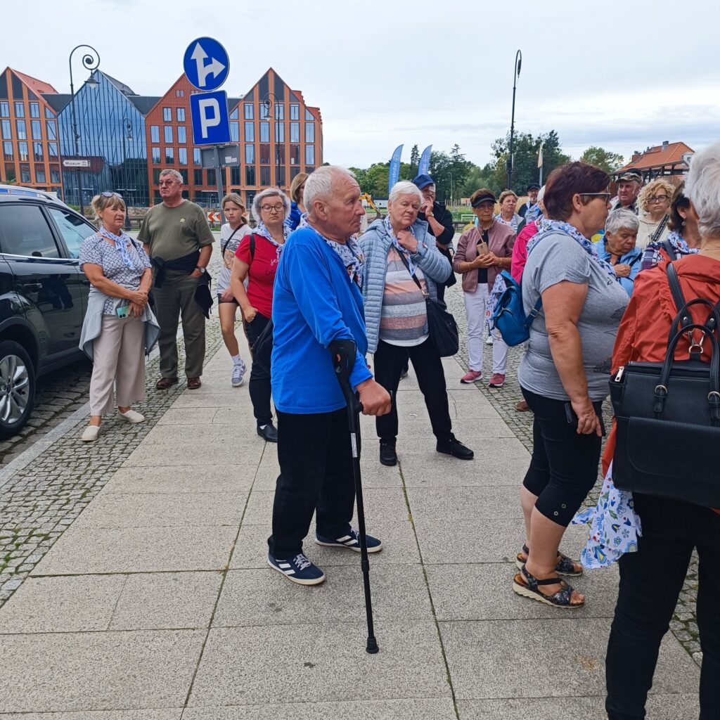 Grupa starszych uczestników wyjazdu rehabilitacyjnego stoi na chodniku w miejskim otoczeniu. Niektórzy używają lasek. Osoby ubrane w codzienne stroje, w tle nowoczesny budynek ze szklaną fasadą, ceglane kamienice z dachami dwuspadowymi oraz zaparkowane samochody. Widoczny znak parkingowy z literą „P” i strzałką w lewo. Atmosfera spacerowa i integracyjna.