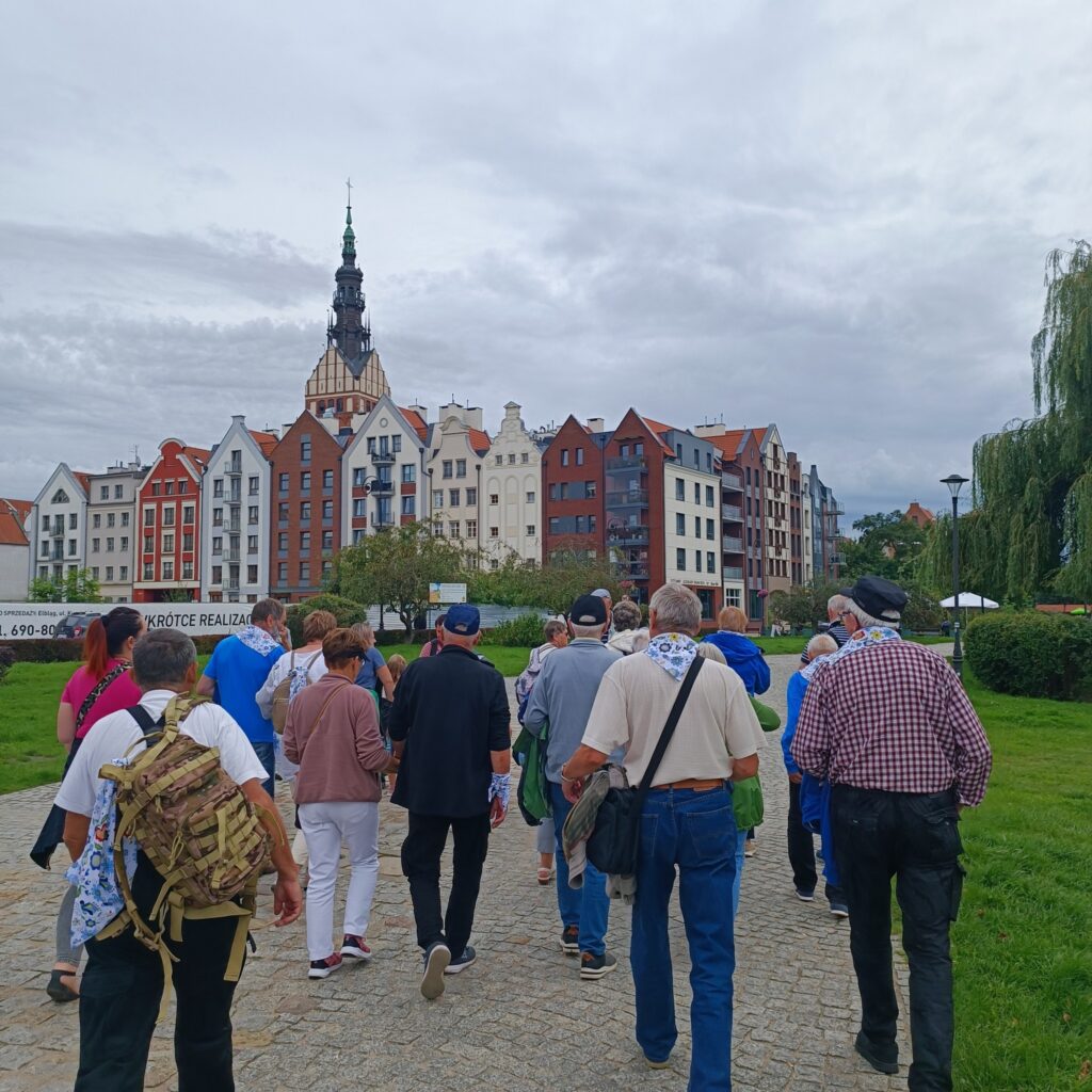 Grupa uczestników wyjazdu rehabilitacyjnego spaceruje brukowaną ścieżką wśród kolorowych, zabytkowych kamienic z ozdobnymi fasadami i spiczastymi dachami. W tle widoczna wieża kościelna. Wiele osób ma niebiesko-białe apaszki. Pogoda pochmurna, otoczenie zielone z drzewami i trawą. Widoczny także szyld z napisem „KROTEC REALIZAC...” przy budynku.