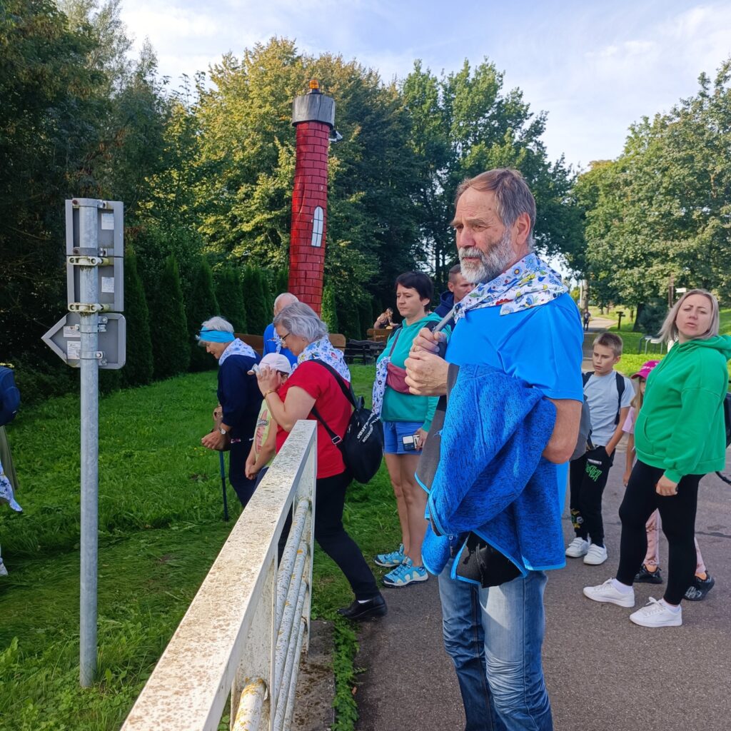 Grupa uczestników wyjazdu rehabilitacyjnego stoi na utwardzonej ścieżce obok białej metalowej barierki, prawdopodobnie na moście. Osoby ubrane w codzienne stroje, wiele z nich ma niebiesko-białe apaszki. Jeden mężczyzna z aparatem i kurtką w ręku stoi na pierwszym planie. W tle zielona trawa, drzewa i czerwona wieża z czarnym szczytem. Widoczny także słupek z oznaczeniami.