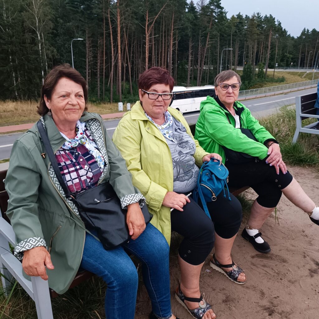 Trzy starsze osoby siedzą na ławce przy drodze, w otoczeniu lasu sosnowego. Ubrane w kurtki, co sugeruje chłodniejszą pogodę. Jedna osoba ma zieloną kurtkę i torbę na ramię, druga jasną kurtkę i okulary, trzecia jaskrawozieloną kurtkę, czarne spodnie i trzyma niebieską torbę. W tle zaparkowany biały autobus i drzewa. Atmosfera spokojna, wypoczynkowa.