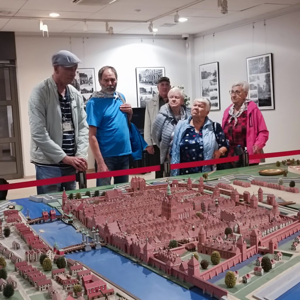 Grupa siedmiu starszych uczestników wyjazdu rehabilitacyjnego stoi za szczegółową makietą historycznego miasta z czerwonymi dachami, kanałami i murami obronnymi. W centrum widoczna duża budowla przypominająca ratusz lub katedrę. Otoczenie muzealne z oświetleniem sufitowym i zdjęciami na ścianach. Atmosfera edukacyjna i skupiona.