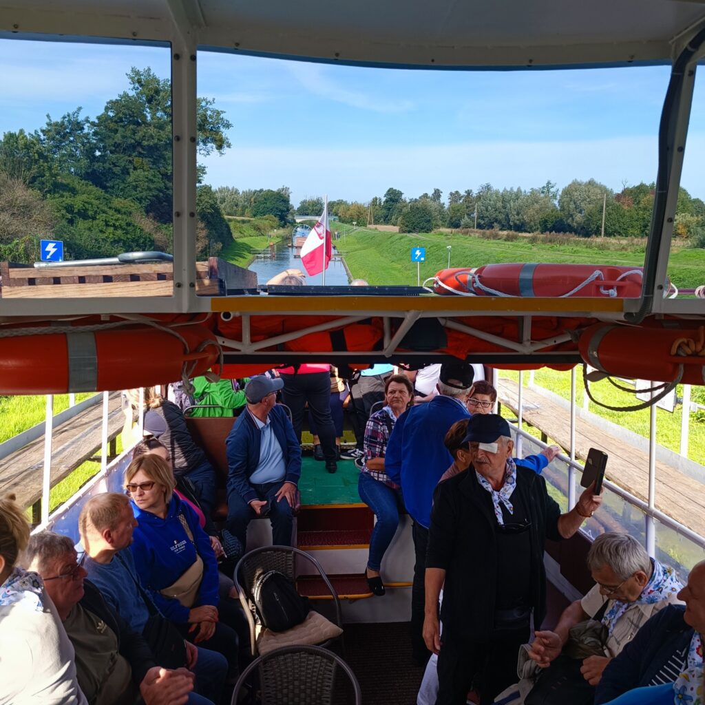 Uczestnicy wyjazdu rehabilitacyjnego płyną łodzią po trawie wzdłuż Kanału Elbląskiego. W tle zielone pola i drzewa, na dziobie widoczna flaga Polski. Na pokładzie znajdują się koła ratunkowe i miejsca siedzące. Nietypowy moment transportu wodnego po lądzie, ukazujący wyjątkową infrastrukturę.