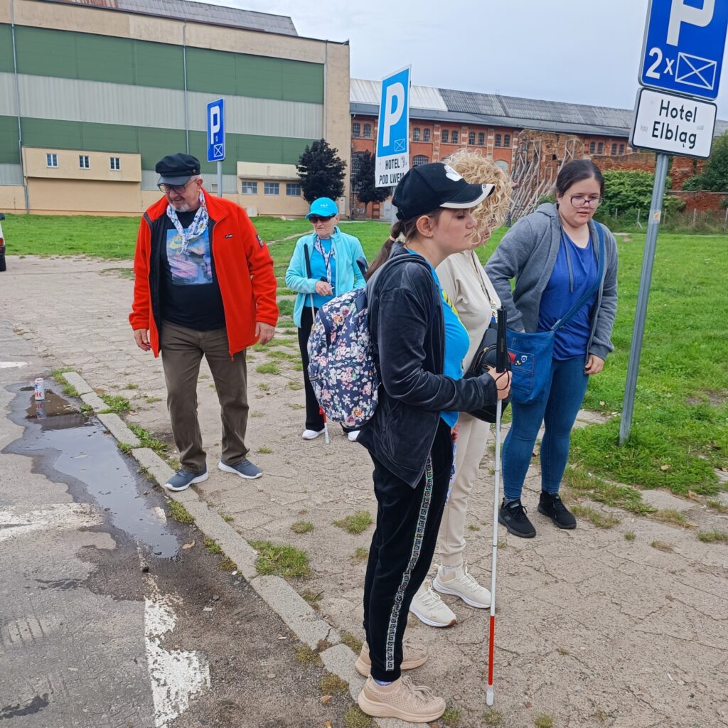 Grupa pięciu uczestników wyjazdu rehabilitacyjnego stoi na utwardzonym placu obok parkingu. Jedna osoba z białą laską i kwiecistym plecakiem jest wspierana przez drugą w białym stroju. Pozostali ubrani w kurtki i czapki. W tle widoczne oznaczenia parkingowe, w tym miejsce dla osób z niepełnosprawnościami oraz szyld „Hotel Elbląg”. Otoczenie miejskie z budynkami i zielenią.
