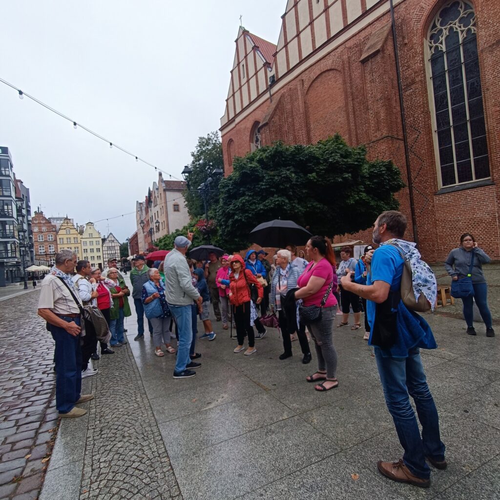 Grupa uczestników wyjazdu rehabilitacyjnego stoi na brukowanej ulicy przed dużym ceglanym budynkiem w stylu gotyckim. Osoby słuchają przewodnika, niektórzy trzymają parasole. W tle wysokie okna z łukami, strome dachy i zawieszone girlandy świetlne. Atmosfera edukacyjna i klimatyczna.