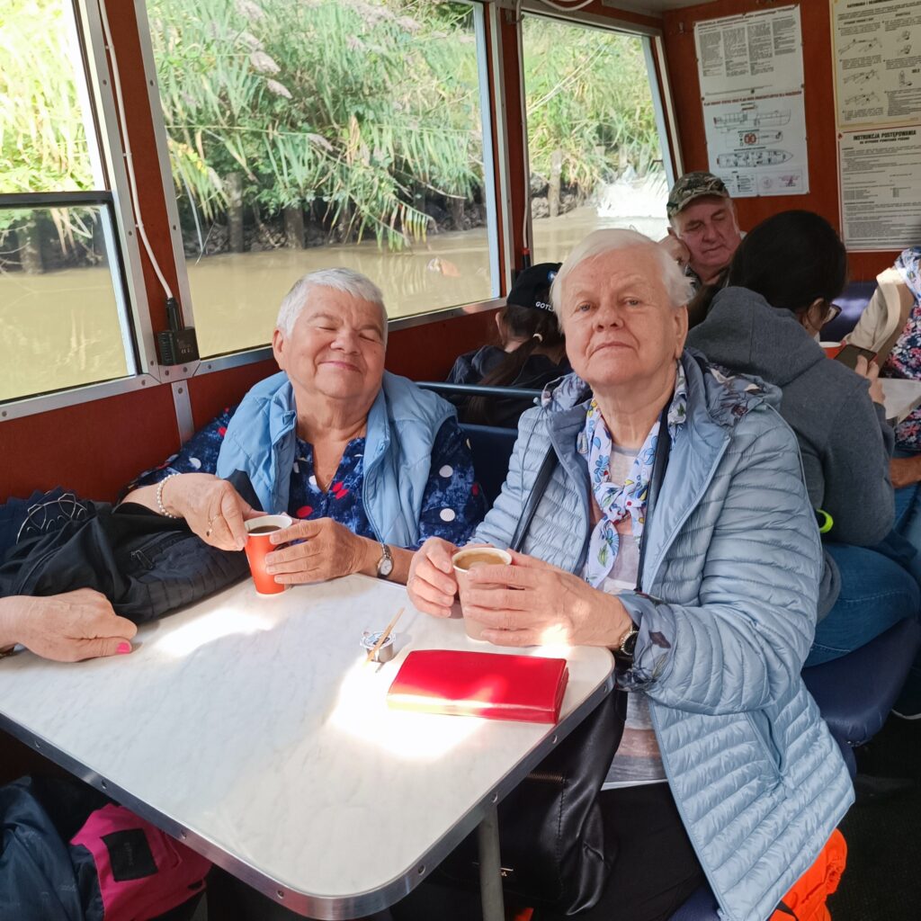 Dwoje starszych uczestników wyjazdu rehabilitacyjnego siedzi przy stoliku wewnątrz statku. Oboje uśmiechnięci, trzymają kubki z napojami. Jedna osoba ubrana w jasnoniebieską kurtkę i kwiecistą bluzkę, druga w szarym płaszczu z apaszką. Na stole leżą okulary, czerwony notes i torby. W tle widać innych pasażerów oraz zielony krajobraz za oknem.