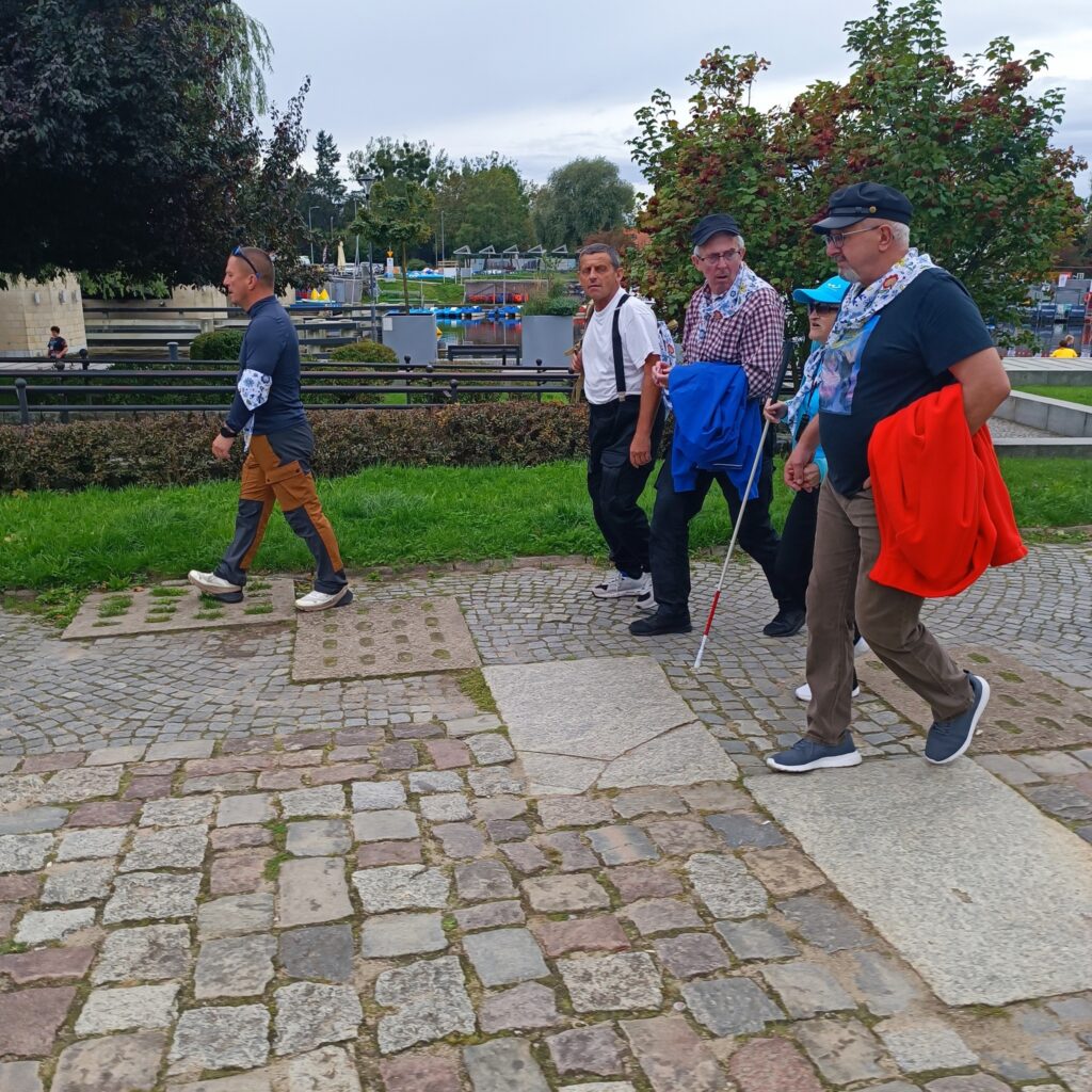Grupa pięciu uczestników wyjazdu rehabilitacyjnego spaceruje po brukowanej i asfaltowej ścieżce w parku lub przestrzeni miejskiej. Jedna osoba używa białej laski, co wskazuje na niepełnosprawność wzroku. W tle widoczne drzewa, trawa, zaparkowane pojazdy i budynki. Atmosfera aktywna i wspólnotowa.