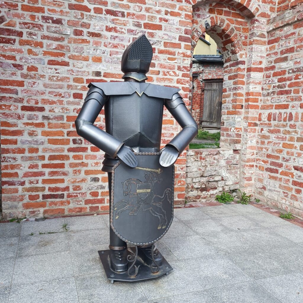 Metalowa rzeźba rycerza stojąca na brukowanym placu przed ceglanym murem z łukowymi przejściami. Rycerz ma hełm z siateczką, zbroję i tarczę z wygrawerowanym koniem oraz zamkiem. Otoczenie sugeruje miejsce historyczne lub wystawę plenerową.