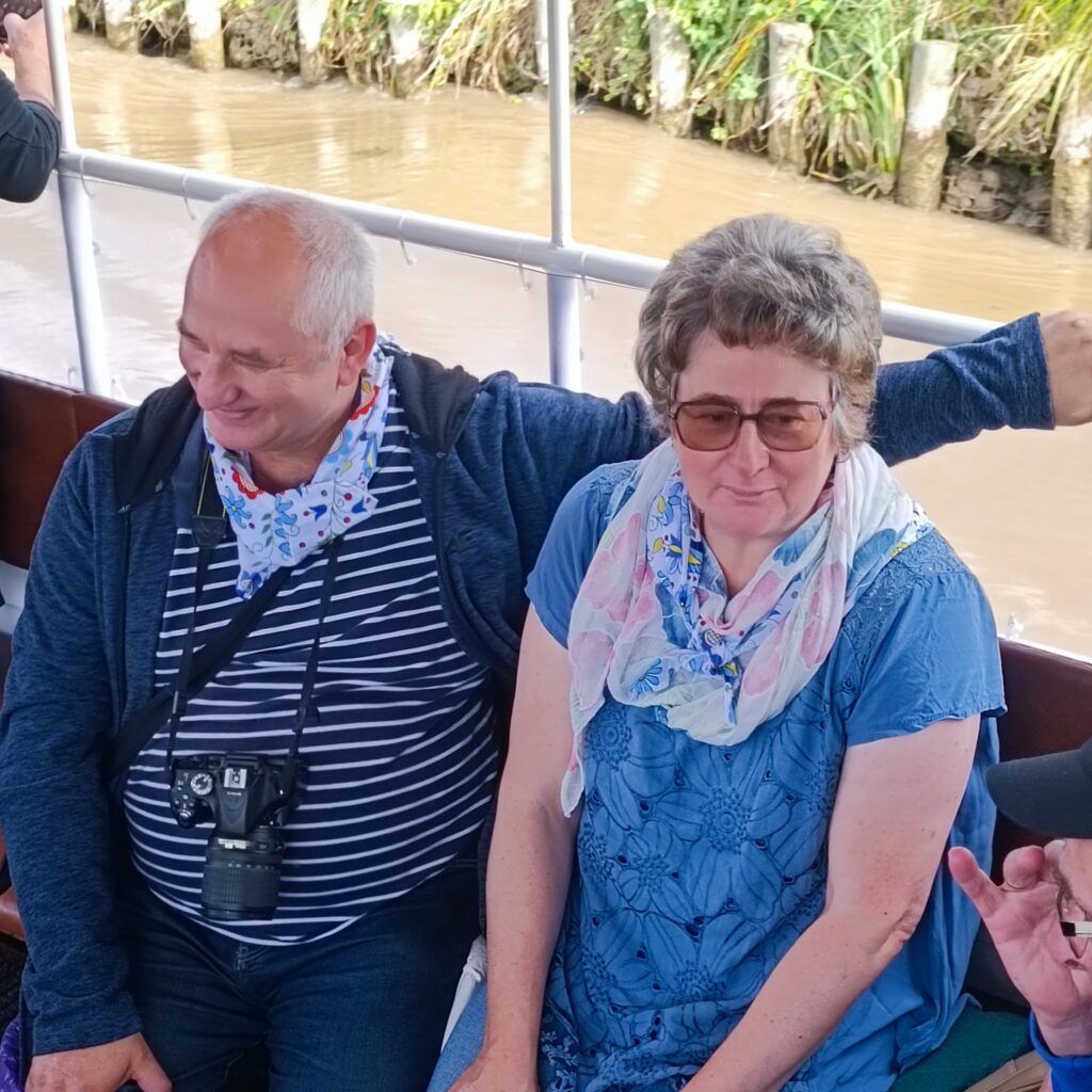 Dwie osoby siedzą obok siebie na łodzi podczas rejsu. Jedna ubrana w granatową kurtkę ze zwisającym aparatem, druga w błękitnej sukience z haftem i okularami przeciwsłonecznymi. Oboje mają jasne apaszki z kwiatowym wzorem. W tle widoczna rzeka i zielony brzeg. Atmosfera spokojna i towarzyska.