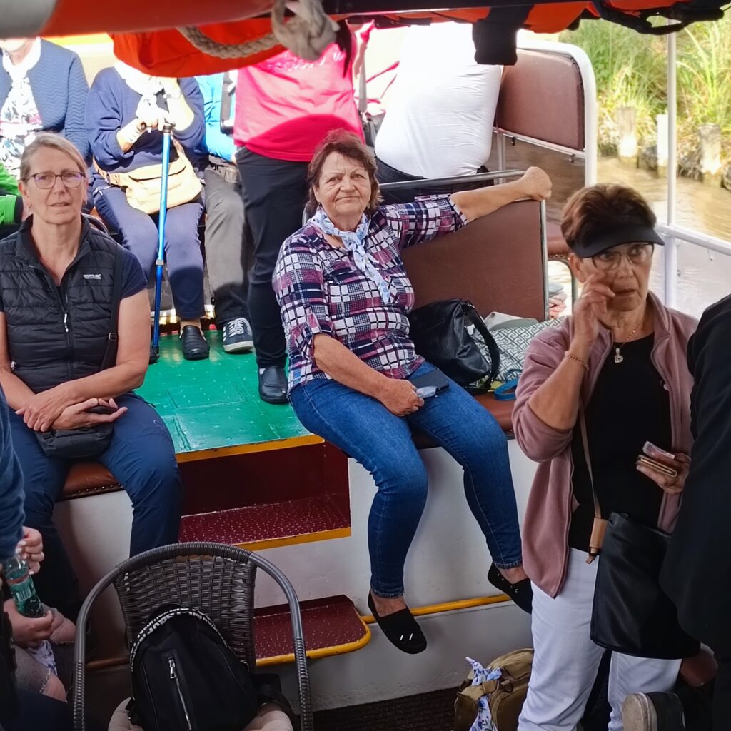 Uczestnicy wyjazdu rehabilitacyjnego siedzą i stoją na górnym pokładzie łodzi z zielono-czerwonymi schodami. Jedna kobieta siedzi na stopniu z ręką opartą o poręcz, inna trzyma butelkę wody. W tle widać trzcinę i wodę. Atmosfera swobodna, pełna rozmów i obserwacji otoczenia.