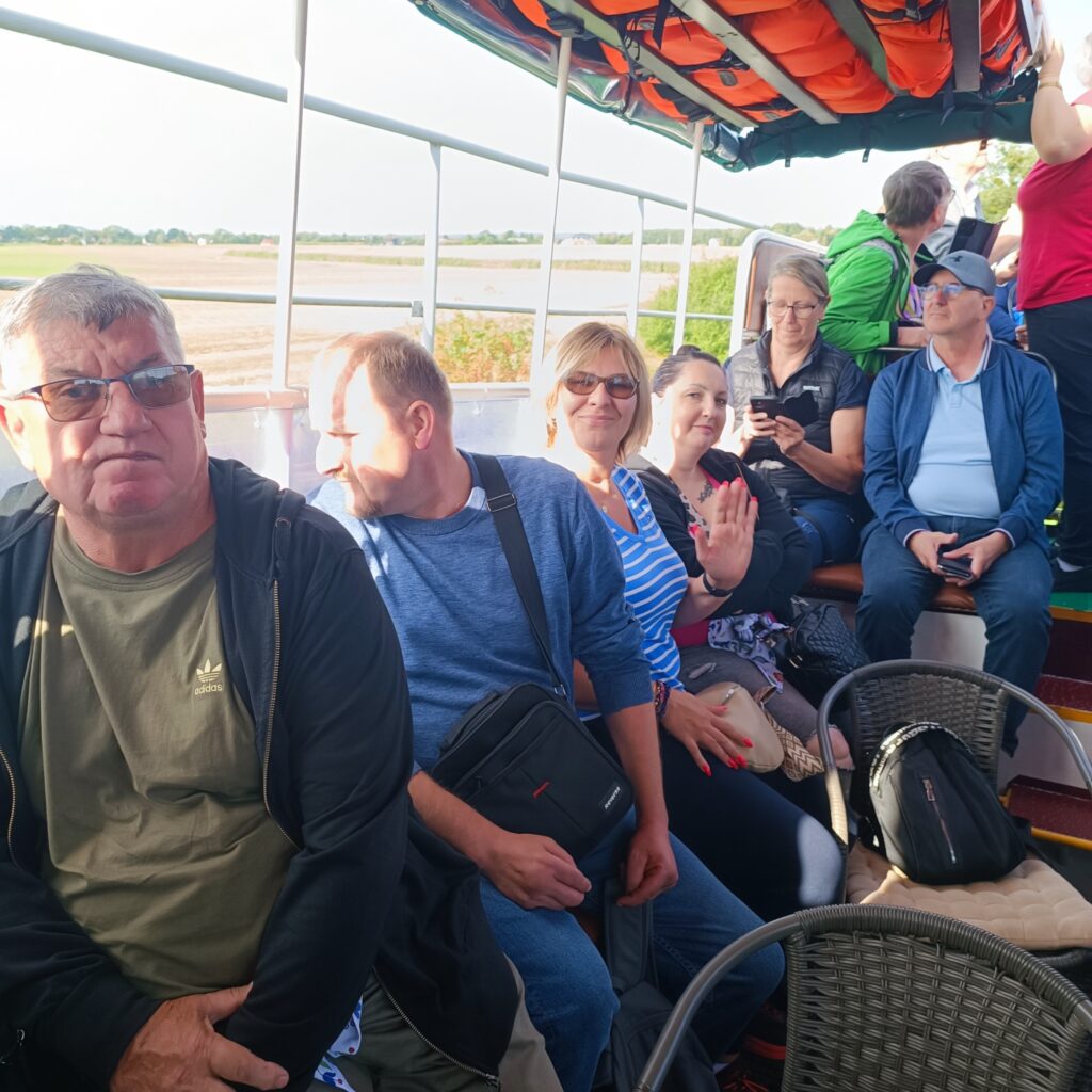 Grupa uczestników wyjazdu rehabilitacyjnego siedzi na ławce pod daszkiem na pokładzie łodzi. Nad głowami widoczne pomarańczowe kamizelki ratunkowe. Część osób patrzy w stronę aparatu, inni korzystają z telefonów lub rozmawiają. W tle płaski krajobraz z polami i drzewami, oświetlony słońcem.