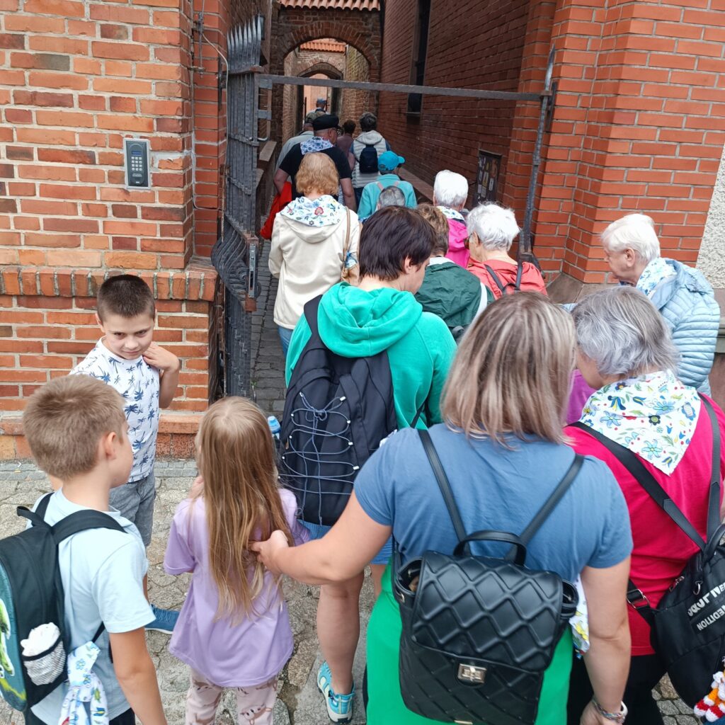 Uczestnicy wyjazdu rehabilitacyjnego, w tym dzieci i dorośli, przechodzą przez wąskie ceglane przejście z łukiem, przypominające zabytkowy portal. Osoby ubrane w codzienne stroje, niektórzy z plecakami. Otoczenie sugeruje historyczne miejsce, np. starówkę lub zamek.