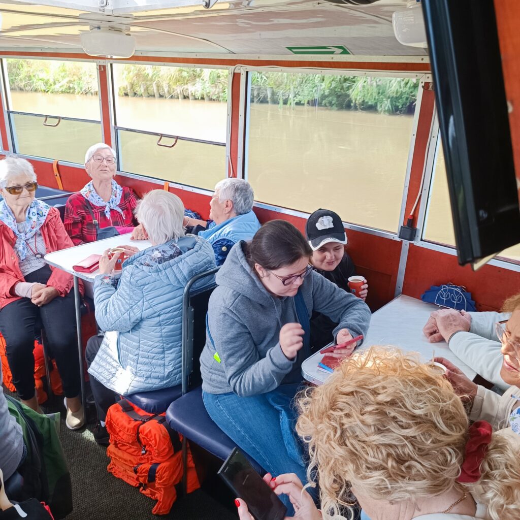 Uczestnicy wyjazdu rehabilitacyjnego siedzą wewnątrz statku z dużymi oknami, przez które widać spokojną rzekę i zielone brzegi. Osoby rozmawiają, jedzą przekąski i odpoczywają przy stolikach z miękkimi siedzeniami. Pod siedzeniami widoczne kamizelki ratunkowe. Atmosfera spokojna i towarzyska.