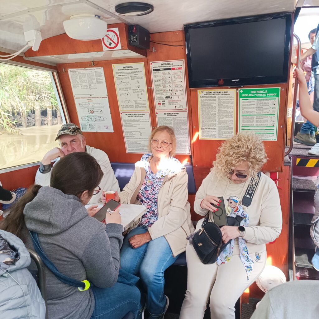 Uczestnicy wyjazdu rehabilitacyjnego siedzą wewnątrz drewnianego statku, rozmawiają, czytają i piją napoje. Jedna osoba pisze na kartce, inni odpoczywają przy oknach z widokiem na wodę i zieleń. Na ścianie widoczne są plakaty informacyjne i telewizor. Jasne światło dzienne wpada przez okna, tworząc przytulną atmosferę.