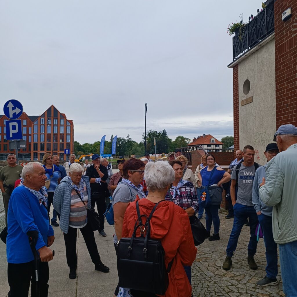 Grupa uczestników wyjazdu rehabilitacyjnego zgromadzona przed budynkiem z napisem „PORT”. Osoby słuchają przewodnika, ubrane w codzienne stroje, niektórzy z plecakami i torbami. W tle nowoczesne budynki, znaki drogowe i flagi. Pogoda pochmurna, otoczenie miejskie z elementami zieleni.