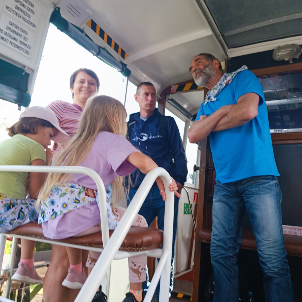 Grupa uczestników wyjazdu rehabilitacyjnego wewnątrz pojazdu — prawdopodobnie autobusu lub tramwaju. Dwoje dzieci siedzi na metalowej ławce, a troje dorosłych stoi obok, rozmawiając i obserwując otoczenie. W tle widoczne są oznaczenia bezpieczeństwa, gaśnica i panel sterowania. Atmosfera swobodna i towarzyska.