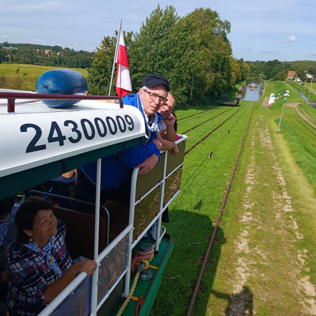 Uczestnicy wyjazdu rehabilitacyjnego jadą otwartą kolejką po trawie wzdłuż Kanału Elbląskiego. W tle widoczna woda, zielone pola i drzewa. Na pojeździe zamontowana flaga Polski. Osoby patrzą na krajobraz, ciesząc się słoneczną pogodą i nietypową atrakcją.