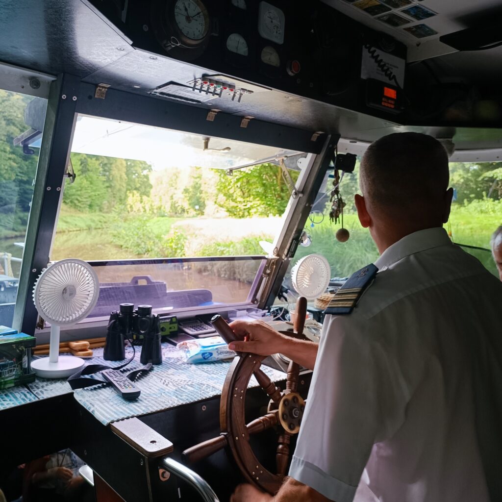 Kapitan w białym mundurze steruje statkiem na Kanale Elbląskim, trzymając drewniane koło sterowe. Wnętrze kokpitu pełne jest przyrządów nawigacyjnych, wentylatorów, lornetki i map. Za oknem widoczna zielona, spokojna sceneria z drzewami i wodą.