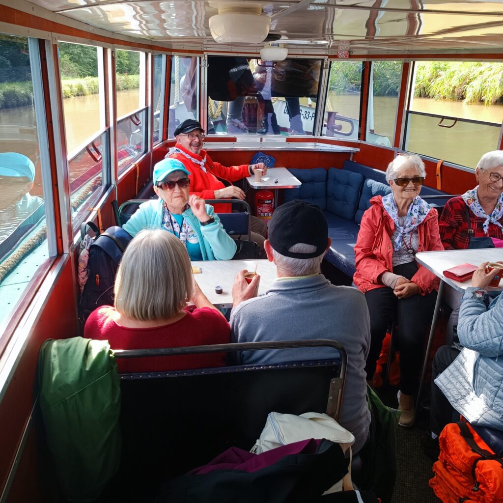Uczestnicy wyjazdu rehabilitacyjnego siedzą wewnątrz przeszklonego statku podczas rejsu po Kanale Elbląskim. W tle widoczne zielone brzegi i spokojna woda. Osoby ubrane w letnie stroje, niektórzy w okularach przeciwsłonecznych i kapeluszach, rozmawiają i odpoczywają przy stolikach. Atmosfera relaksu i wspólnego spędzania czasu.