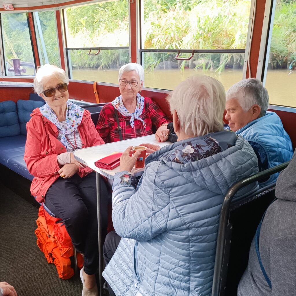 Cztery uczestniczki wyjazdu rehabilitacyjnego odpoczywają podczas rejsu statkiem po Kanale Elbląskim. Siedzą przy stoliku w środku statku, rozmawiają i delektują się napojami. Uśmiechają się, w tle widać zieleń przy brzegu. Uczestniczki są ubrane w kolorowe kurtki i chusty, co dodaje radosnej atmosfery tej chwili.