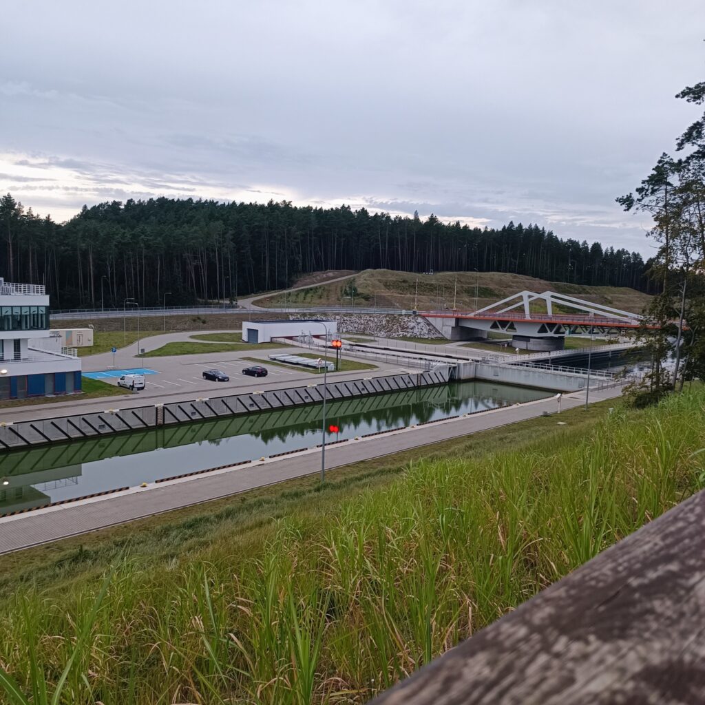 Widok na Przekop Mierzei Wiślanej z nowoczesną infrastrukturą, w tym mostem i kanałem wodnym. W tle widać malowniczy las, a na pierwszym planie znajduje się parking i część urządzeń związanych z przystanią. Całość otoczona jest zielenią, a most na pierwszym planie podkreśla nowoczesny charakter tego miejsca.